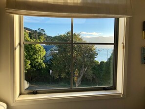 A window with a view of a coastal area, including trees, a house, and boats on the water.