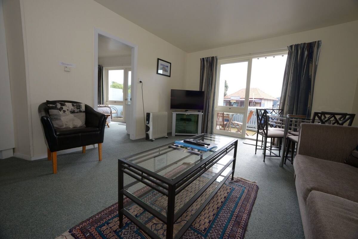 Ein Wohnzimmer mit Glastisch, schwarzem Sofa, beigem Sofa, Fernseher und Blick ins Freie.