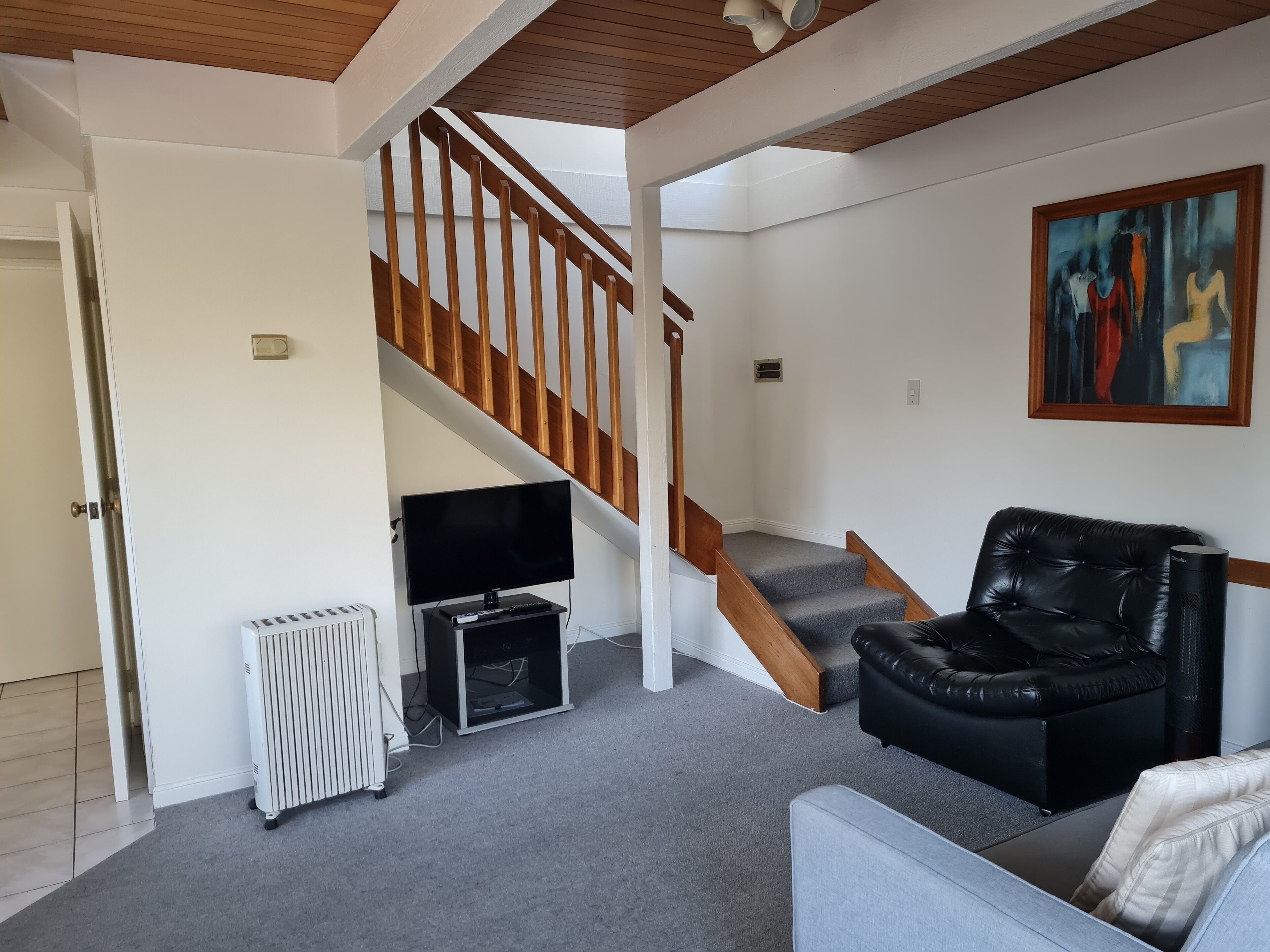 A living room with a staircase, a black leather sofa, a grey sofa, a TV, and a radiator.