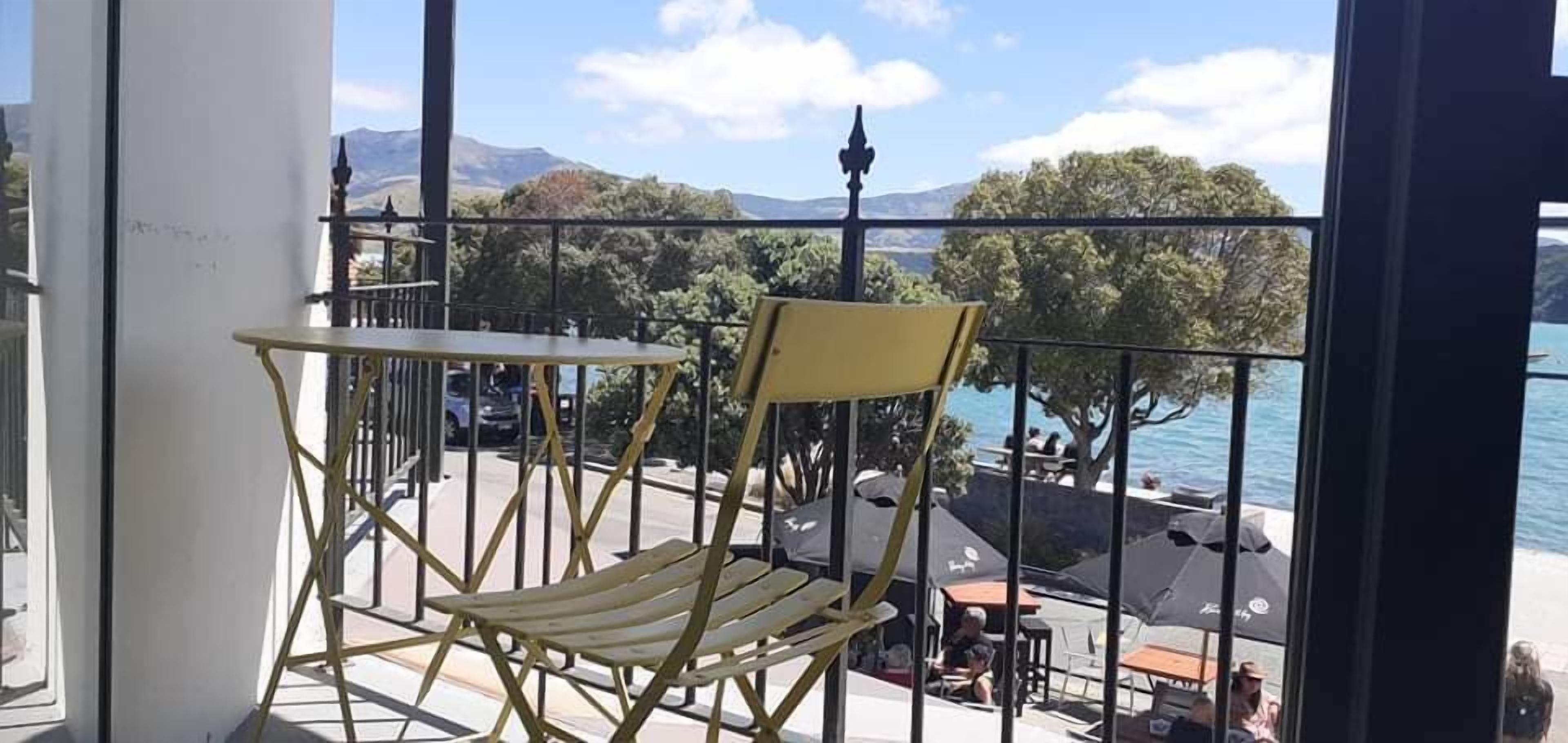 A balcony with a yellow chair and table, overlooking a lakeside area with umbrellas and people.