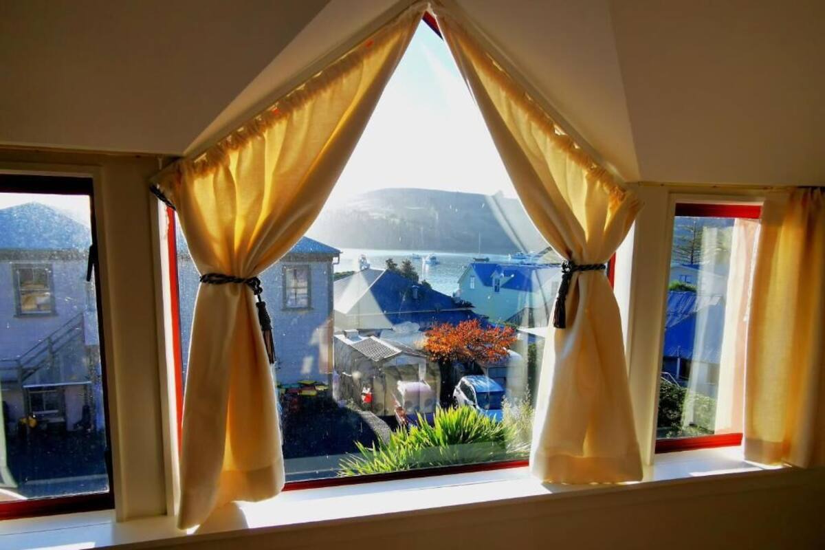 Ein Zimmer mit einem großen Fenster, dessen gelbe Vorhänge zurückgebunden sind und einen Blick auf eine Wohngegend mit Häusern und einem Berg im Hintergrund freigeben.