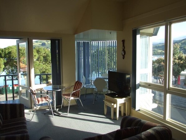 A living room with a TV, a dining table, and chairs. There are large windows with a view of greenery and buildings.