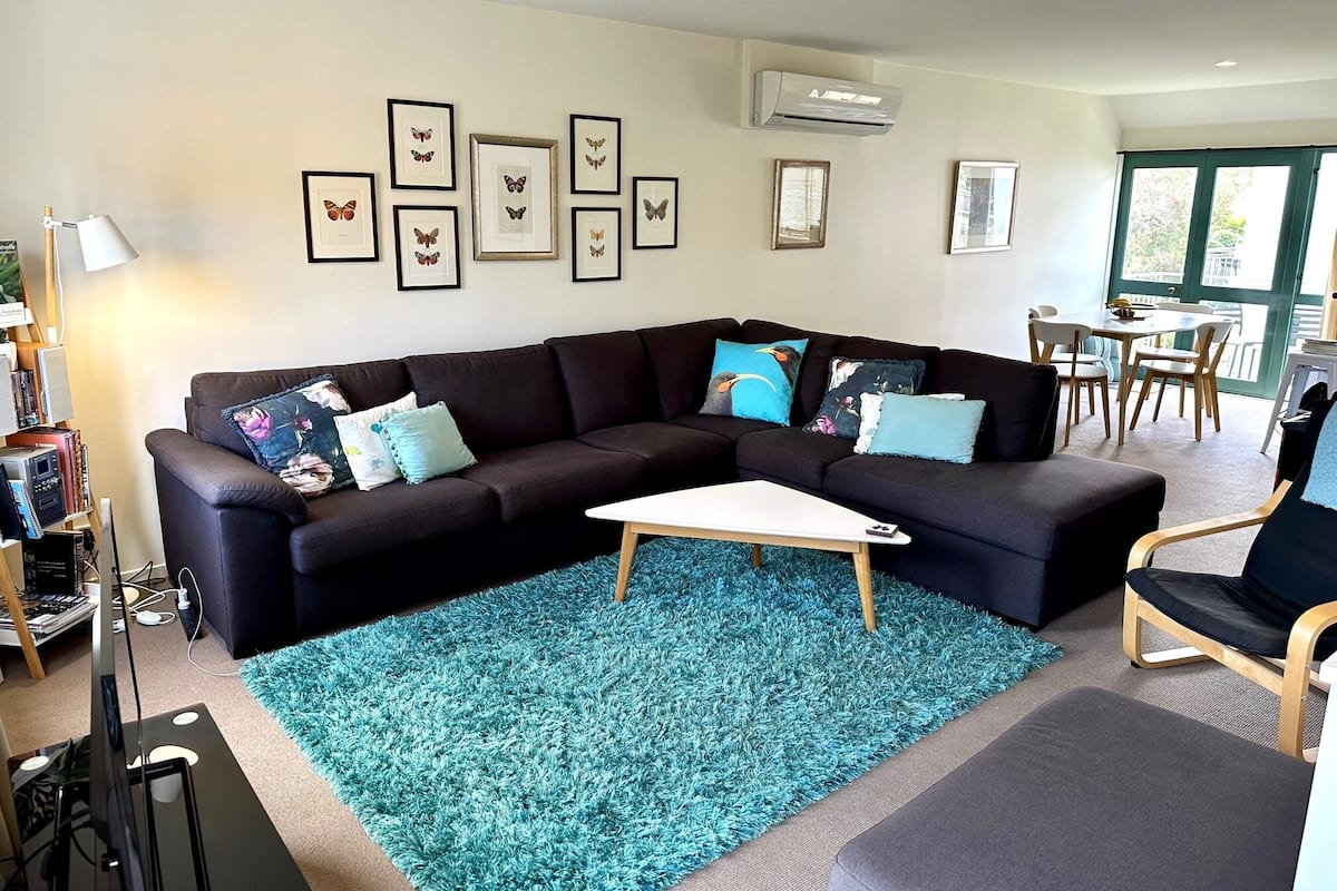 Ein modernes Wohnzimmer mit einem schwarzen Ecksofa, einem türkisfarbenen Teppich und gerahmten Schmetterlingsbildern an der Wand.