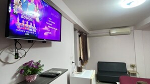 Quarto pequeno e bem iluminado com televisão na parede exibindo um programa. Há uma escrivaninha com cadeira, uma planta pequena e uma cama com cobertor.