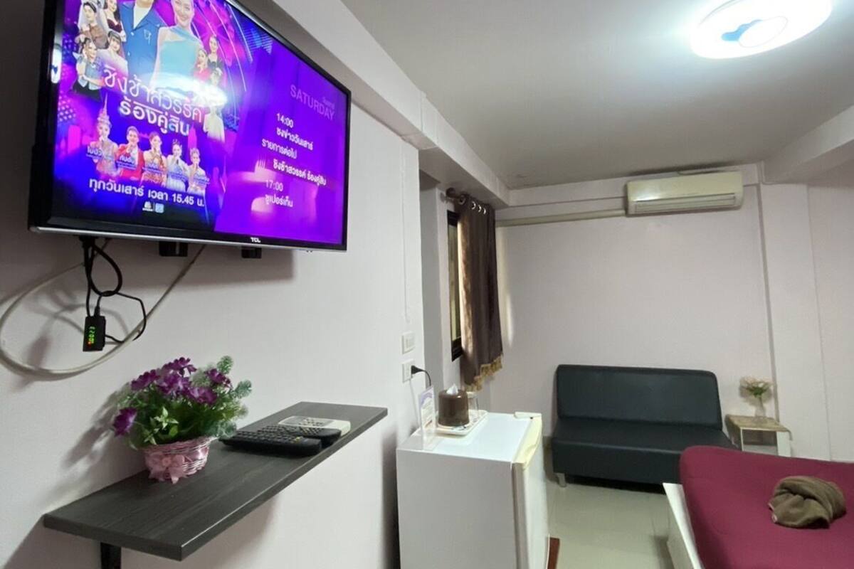A small, well-lit room with a television mounted on the wall displaying a program. There is a desk with a chair, a small plant, and a bed with a blanket.