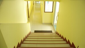 Uma escadaria com degraus de madeira e um tapete preto na base, que leva a um corredor claro com paredes amarelas e uma janela.