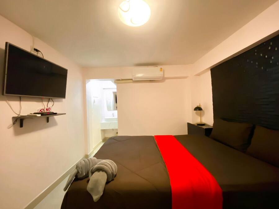 A bedroom with a bed, a TV mounted on the wall, an air conditioning unit, and a nightstand with a lamp.