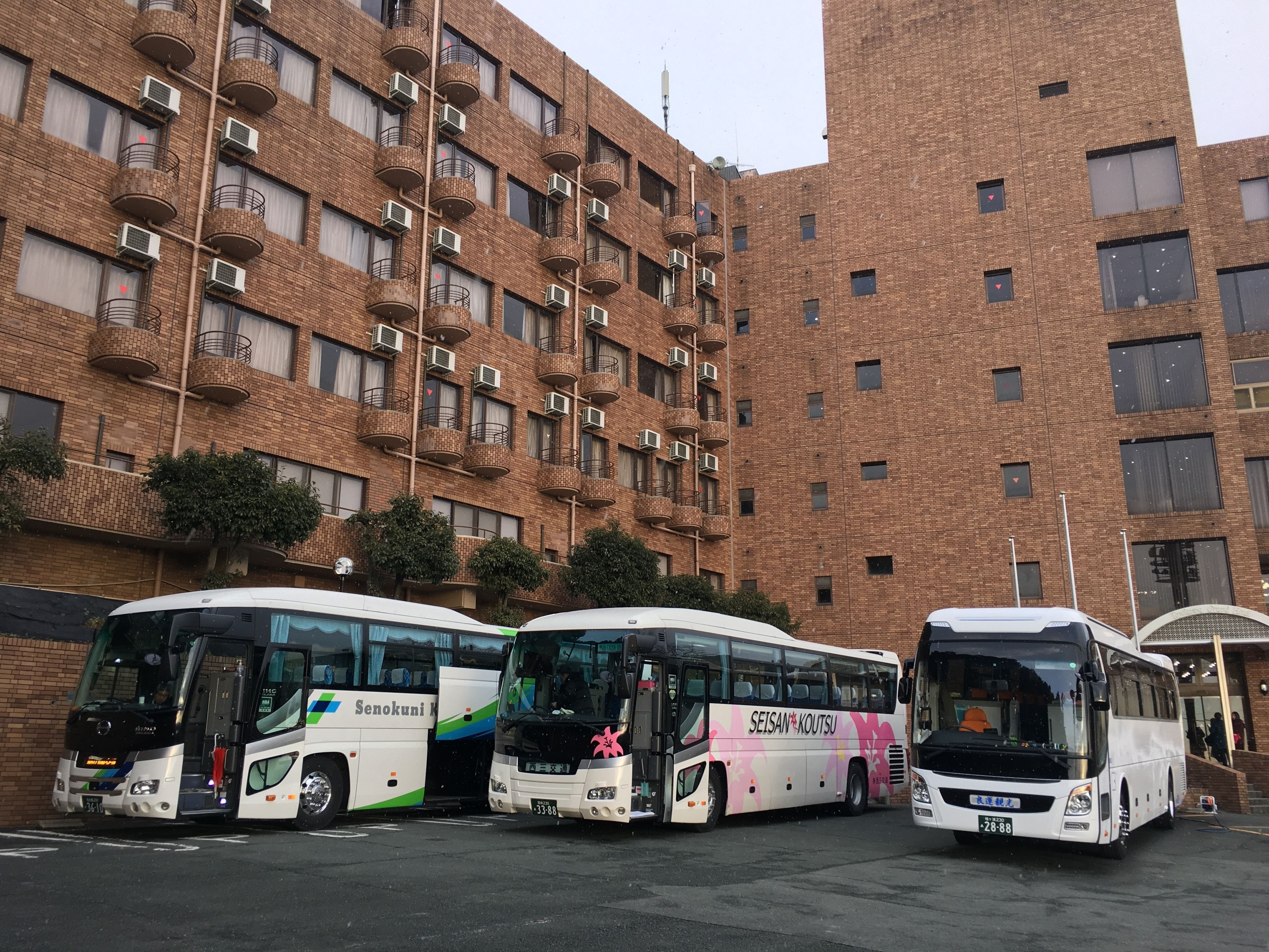 Three buses parked in front of a multi-story brick building with balconies and air conditioning units.