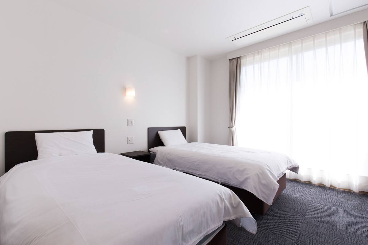 Een hotelkamer met twee bedden, een nachtkastje, een wandlamp en een groot raam met dunne gordijnen.
