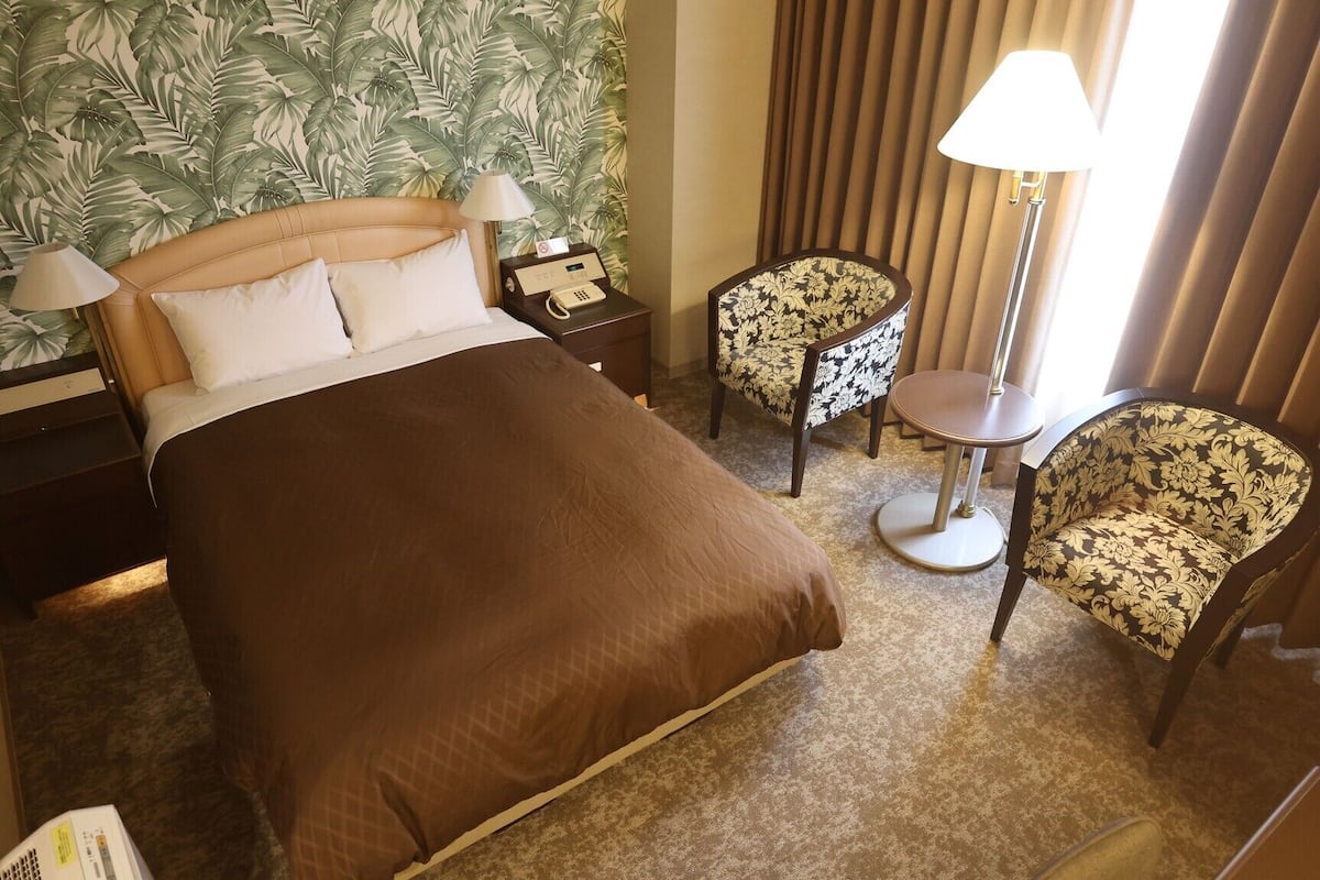 A hotel room with a bed, two chairs, a bedside table, a lamp, and a telephone.