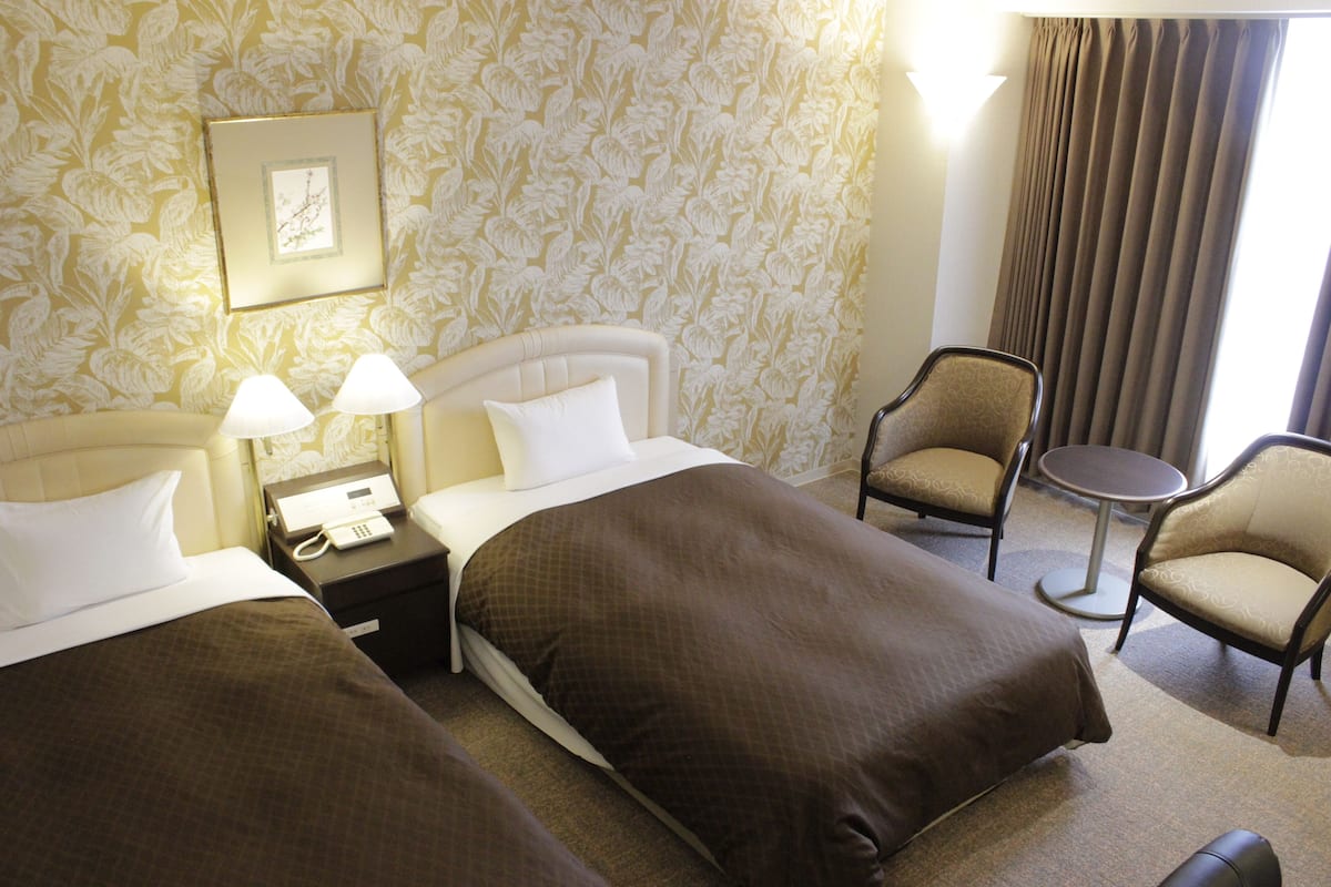 A hotel room with two beds, a bedside table with a phone, two lamps, a chair, and a small round table.