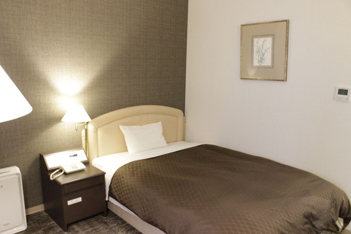 A hotel room with a bed, a bedside table with a lamp, a phone, and a thermostat.