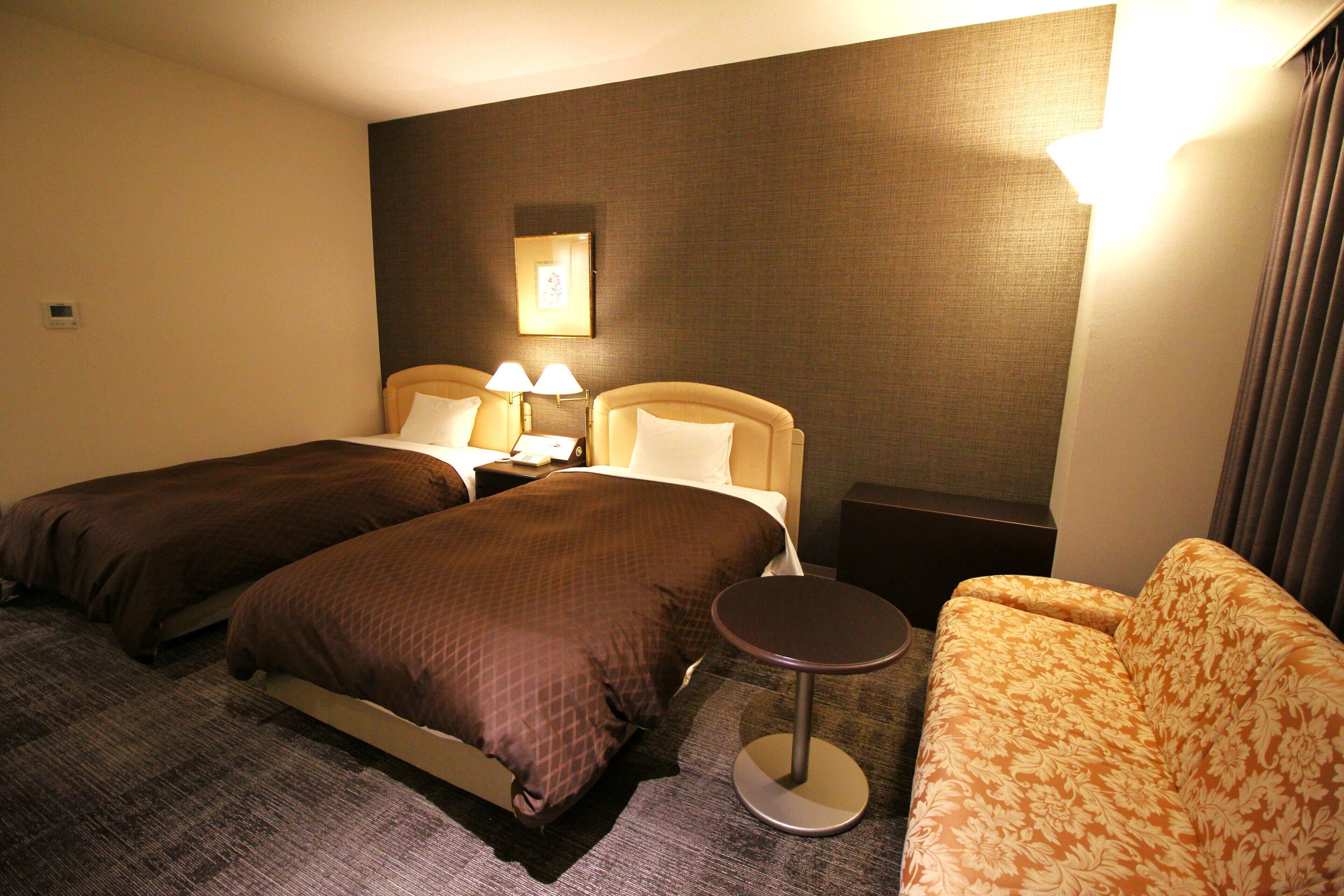 Habitación de hotel con dos camas individuales, una mesita redonda pequeña, un sofá y una mesita de noche con lámparas.