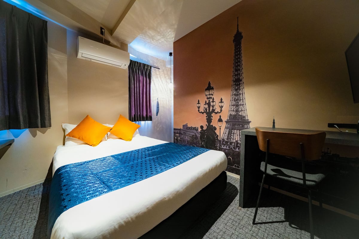 A hotel room with a bed, a desk, a chair, a wall-mounted air conditioner, and a mural of the Eiffel Tower.
