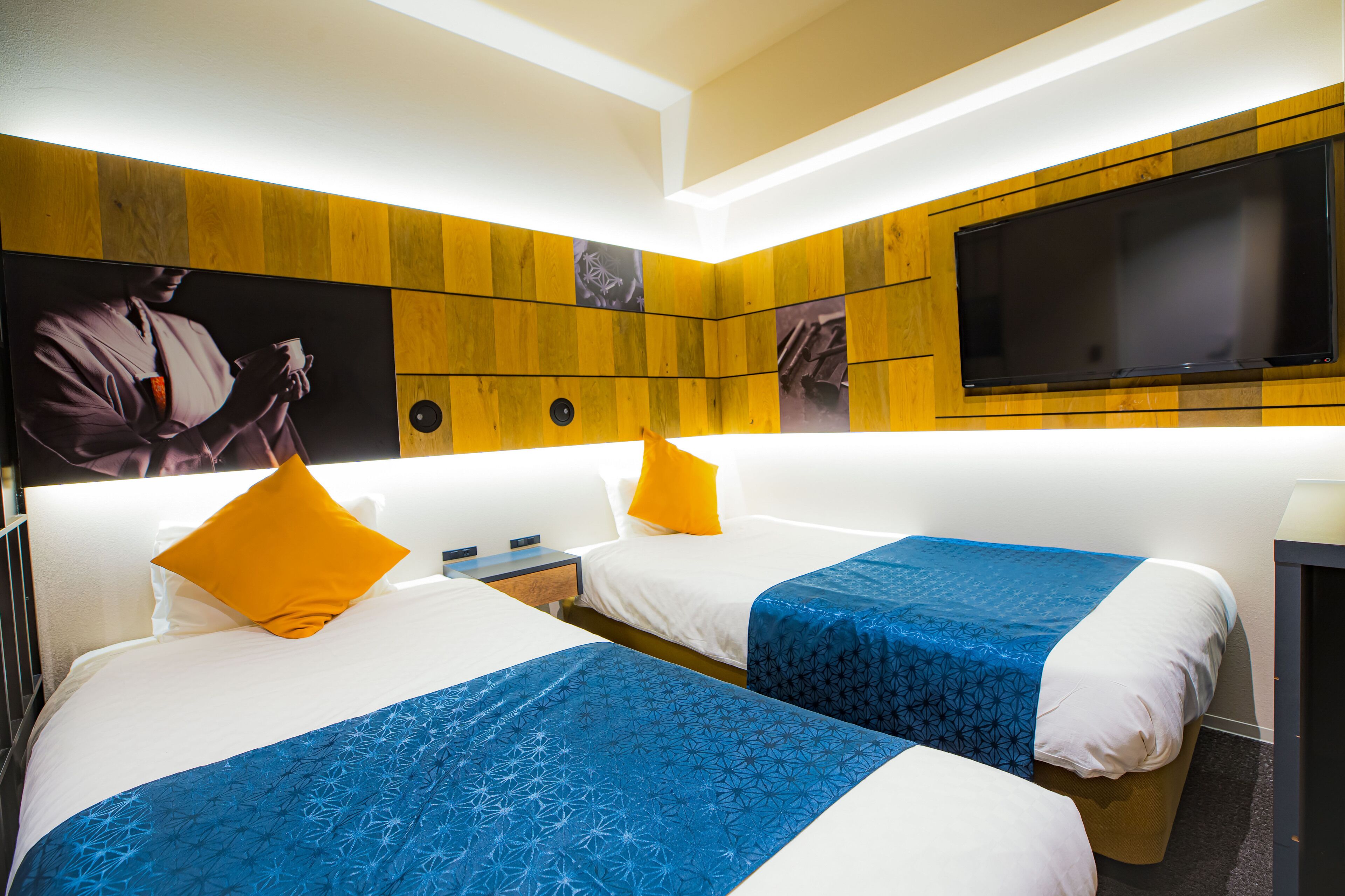 A hotel room with two beds, each with a blue and white bedspread, a large flat-screen TV mounted on the wall, and a wooden panel design on the wall behind the beds.