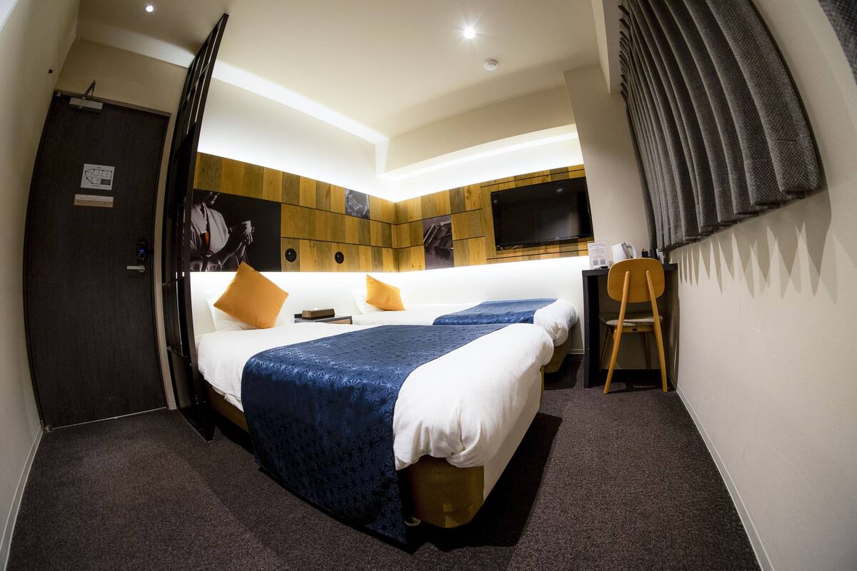 A hotel room with two beds, a desk, a chair, a TV, and a door.