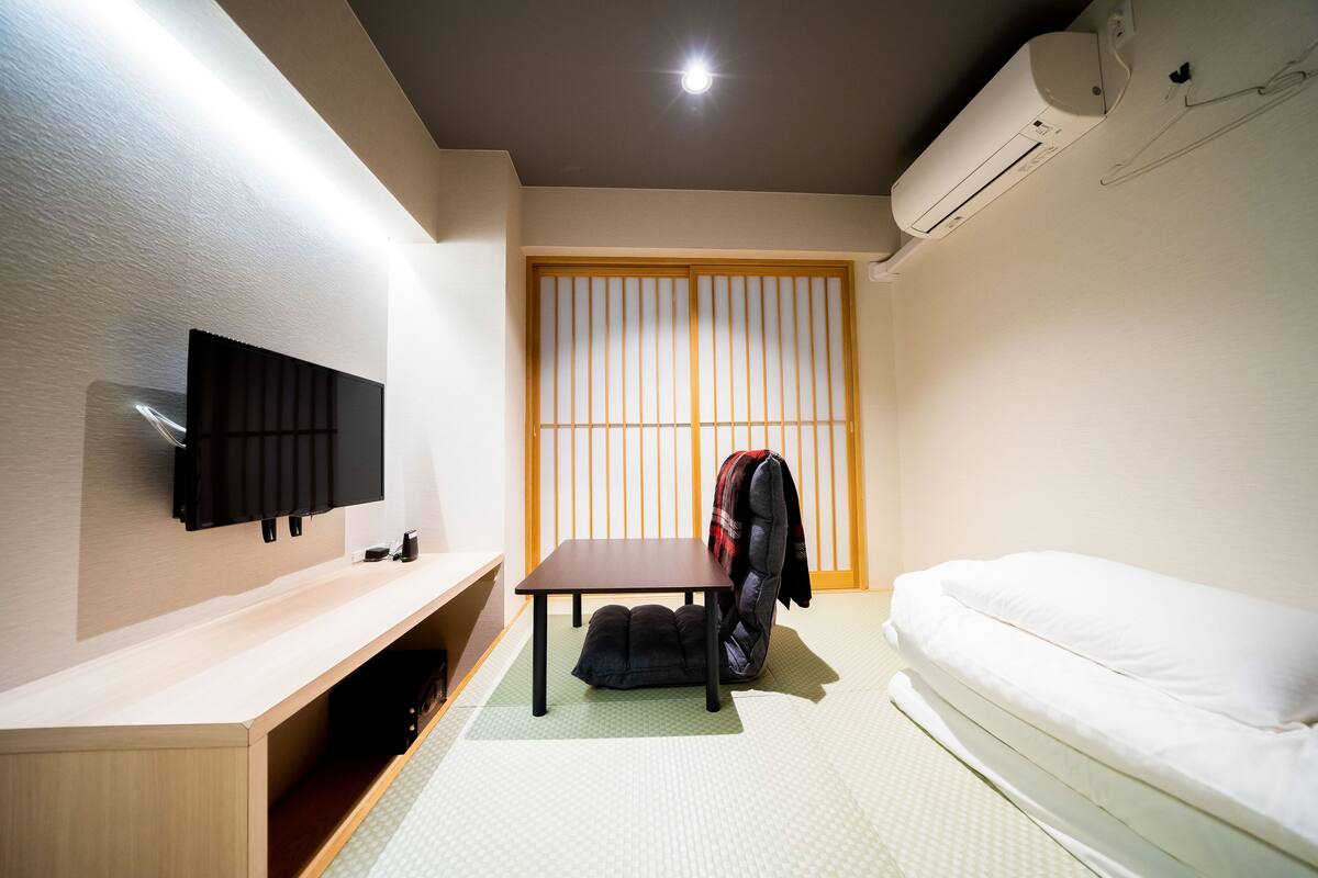 A hotel room with a TV mounted on the wall, a small table, a futon bed, and a sliding shoji screen door.