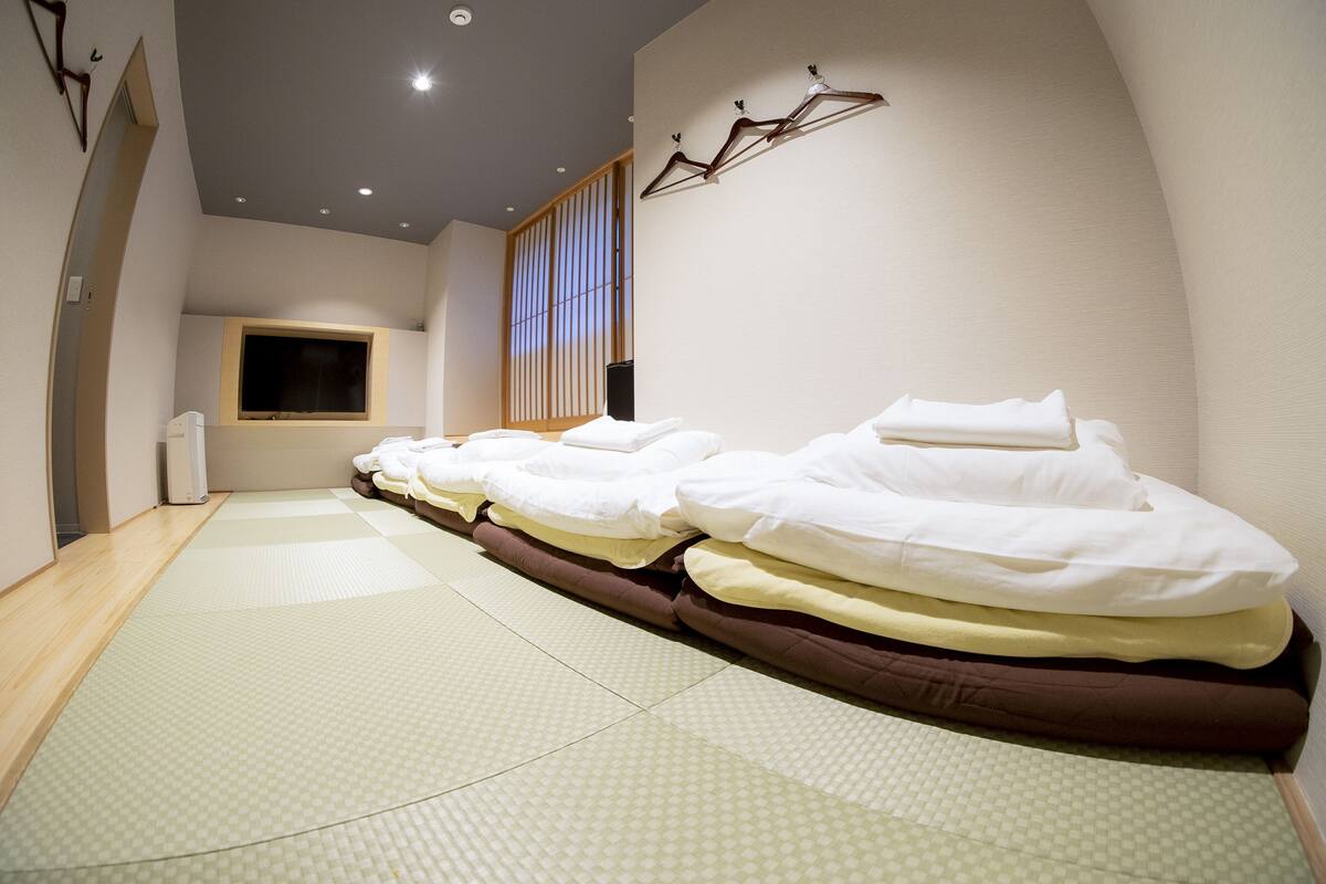 A room with multiple beds aligned along one wall, a large window with vertical blinds, and a wall-mounted heater.