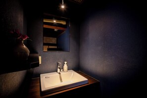 A bathroom with a white sink, a mirror, and a vase with flowers. - Okamotoya Ryokan (Beppu)
