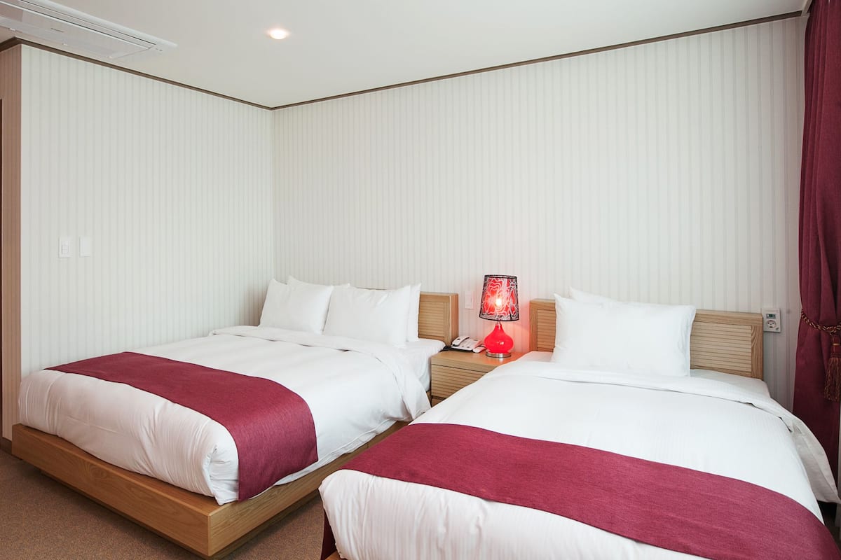 Ein Hotelzimmer mit zwei Betten, jeweils mit weißer Bettwäsche und einer roten Decke. Auf dem Nachttisch zwischen den Betten steht eine rote Lampe.