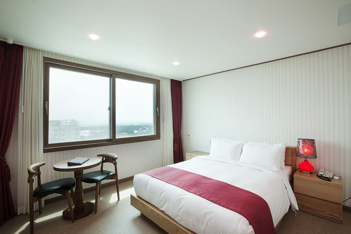 Ein Hotelzimmer mit einem großen Bett, einem Schreibtisch mit Stuhl, einem Nachttisch mit Lampe und einem Fenster mit Vorhängen.