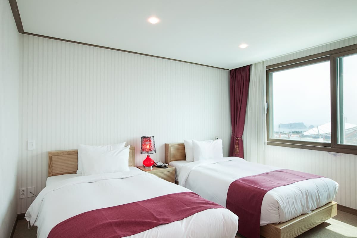 Ein Hotelzimmer mit zwei Betten, einem Nachttisch, einer roten Lampe und einem Fenster mit Blick auf eine entfernte Struktur.