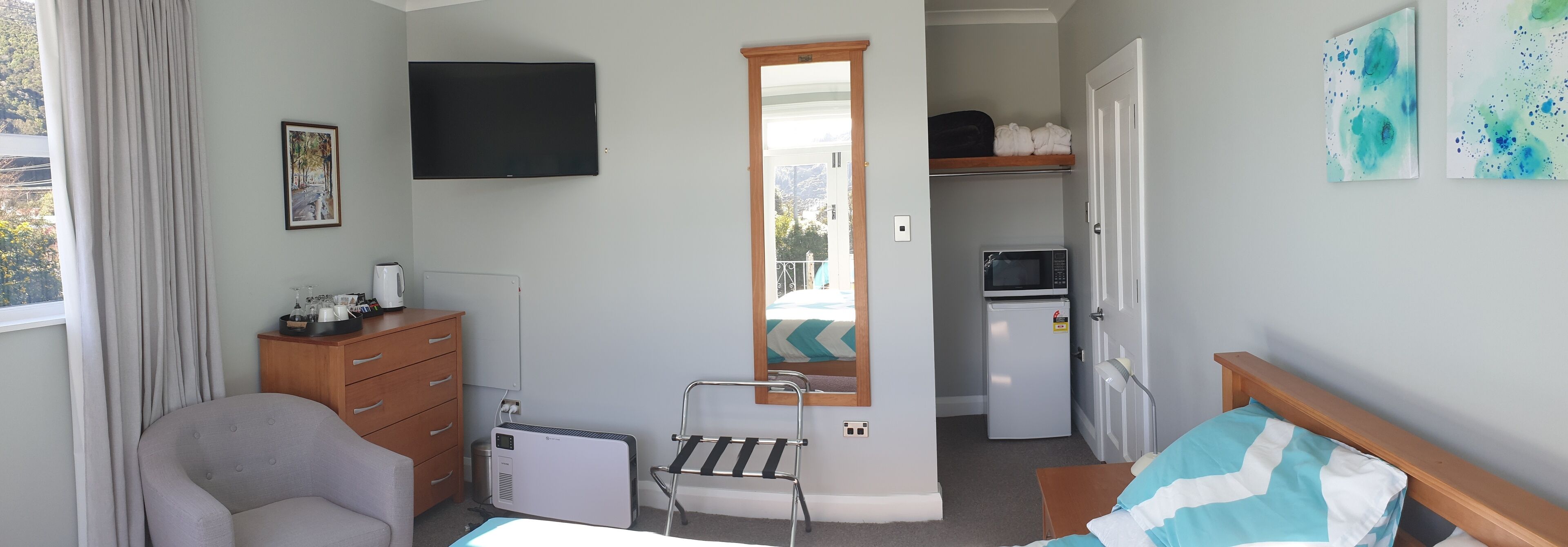 A hotel room with a bed, a desk, a chair, a TV, a small kitchenette, and a view of the outdoors.