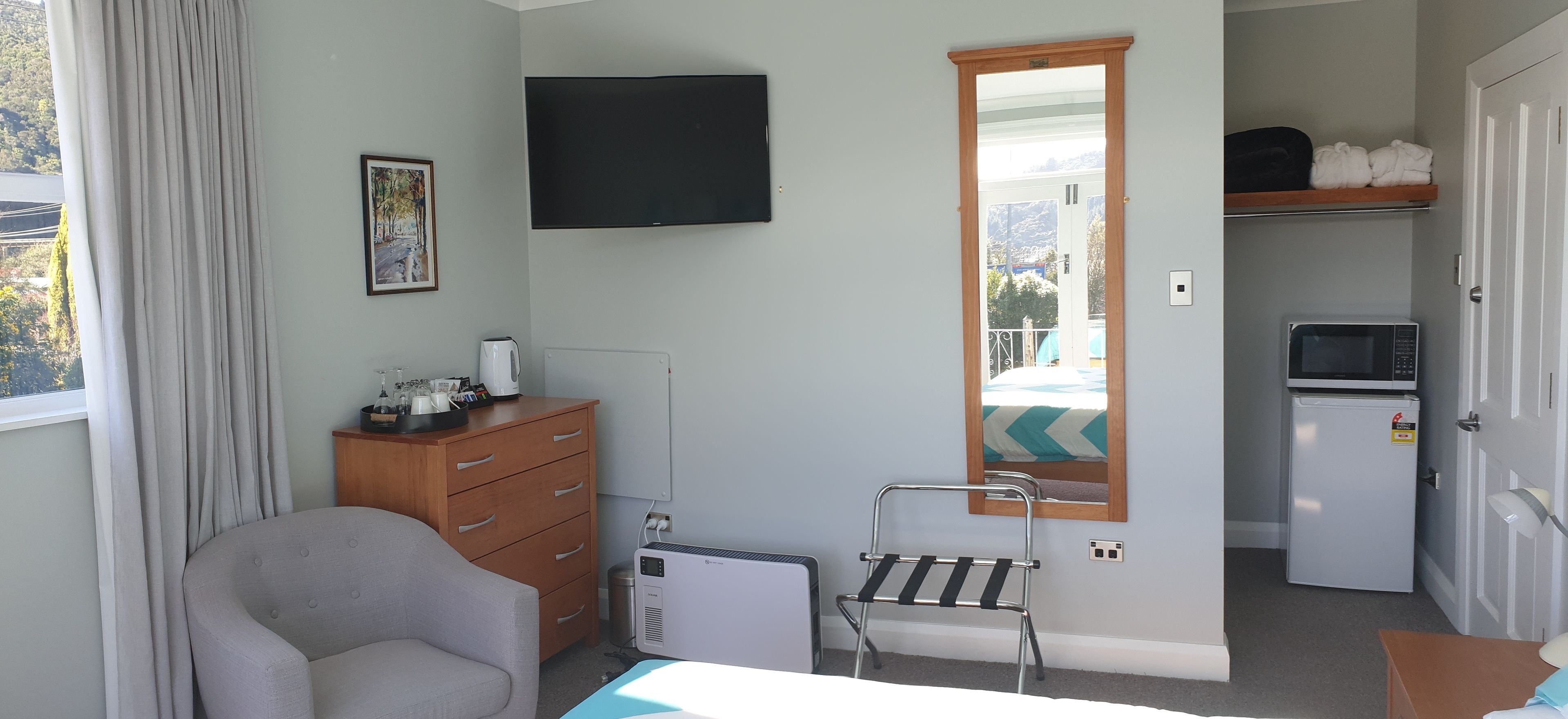 Une chambre d’hôtel comprenant un lit, une chaise, une commode, un téléviseur, un miroir, un radiateur, un four à micro-ondes et une porte donnant sur un balcon.