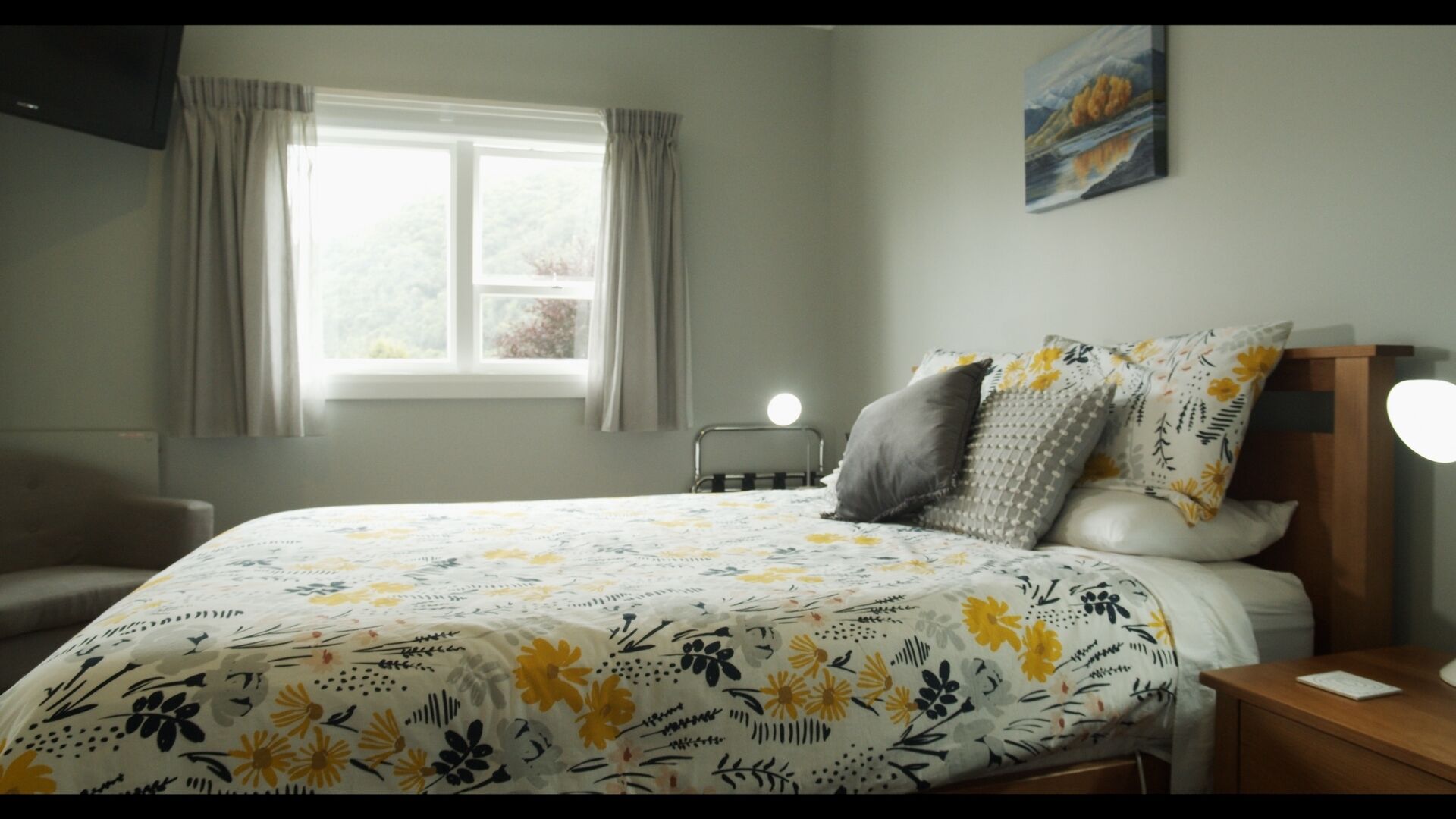 A bedroom with a bed, a bedside table, a nightstand, a window with curtains, and a painting on the wall.