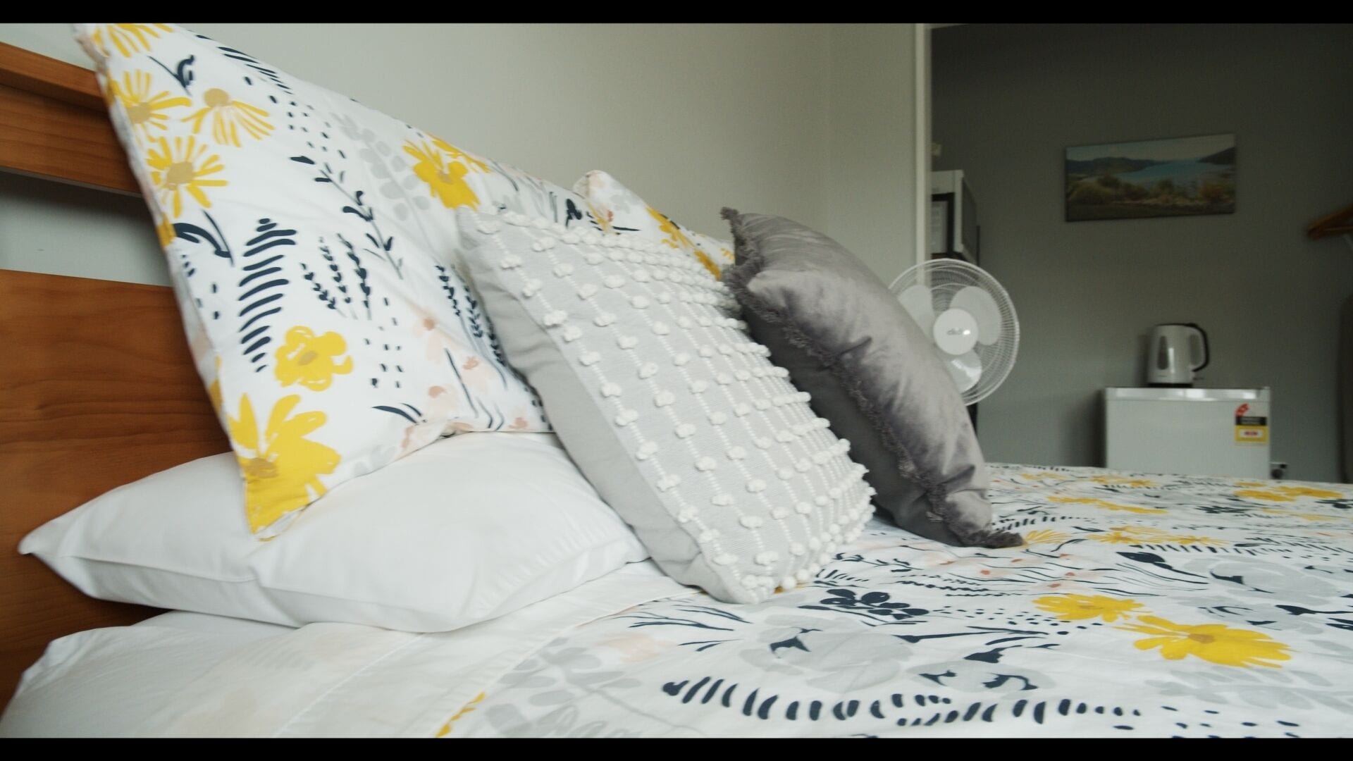 A bed with a floral-patterned duvet, a wooden headboard, and multiple pillows including a white one with a textured design.