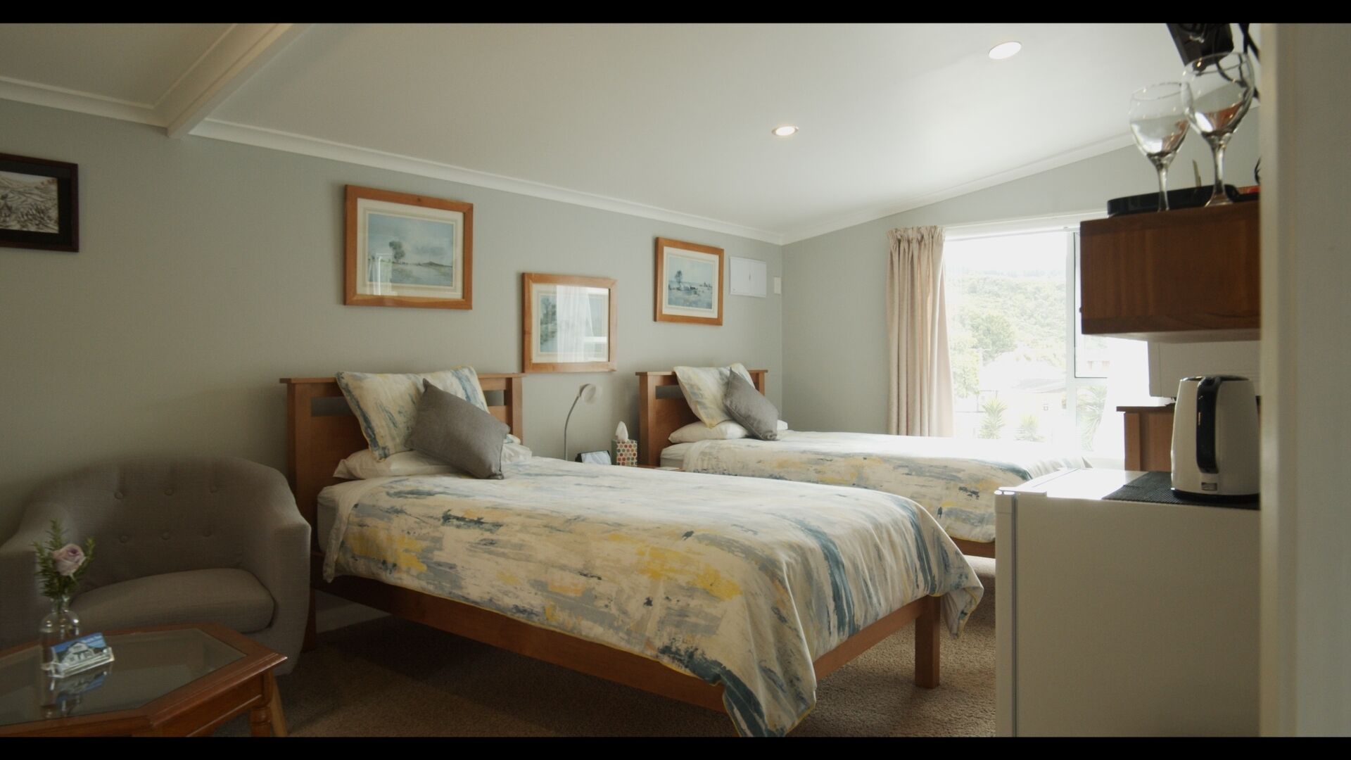 A hotel room with two beds, a sitting area with a chair, a small table, and a window with curtains.