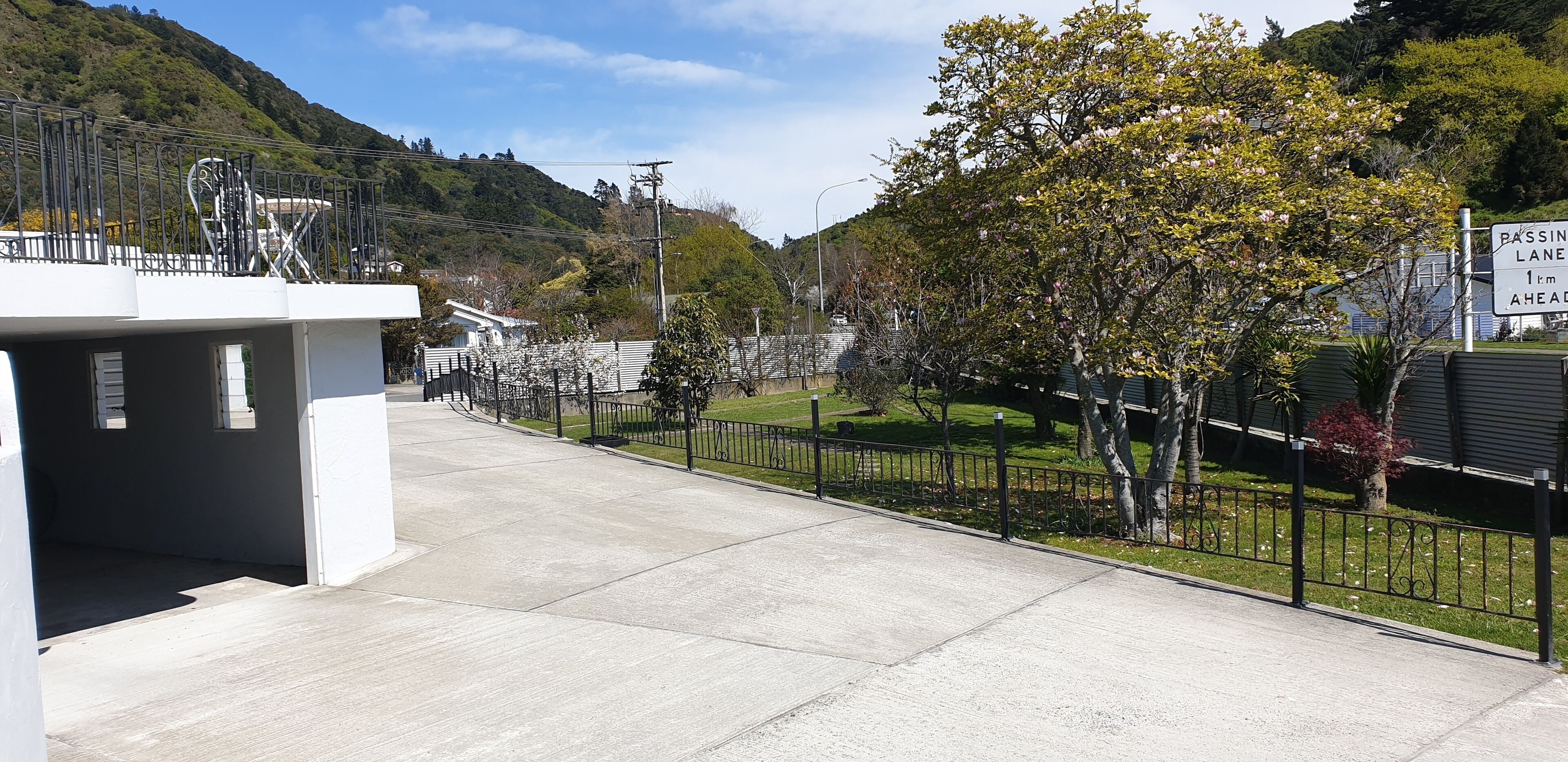 Un sendero para peatones con una cerca de metal, un cartel que indica un carril de adelantamiento a 1 km y un edificio de fachada blanca.
