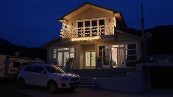 Una casa de dos pisos con un balcón decorado con luces, un coche estacionado frente a ella y una antena parabólica en el techo.
