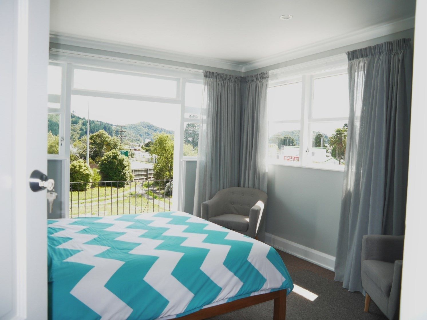 A bedroom with a bed, a chair, and large windows offering a view of greenery and buildings.