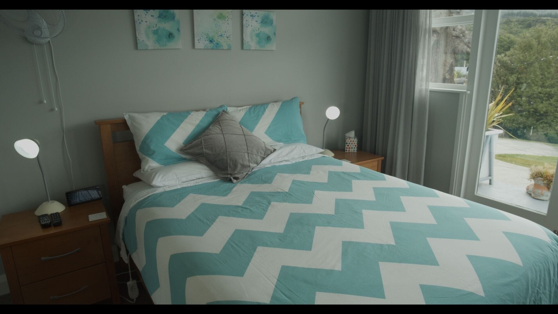 A neatly made bed with a chevron-patterned bedspread, a bedside table with a lamp, and a window with a view of greenery.