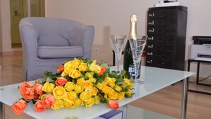 Ett vardagsrum med ett glasbord där en bukett gula och orange rosor står, tillsammans med två champagneglas och en flaska champagne. En grå fåtölj och ett svart skåp med lådor syns också.
