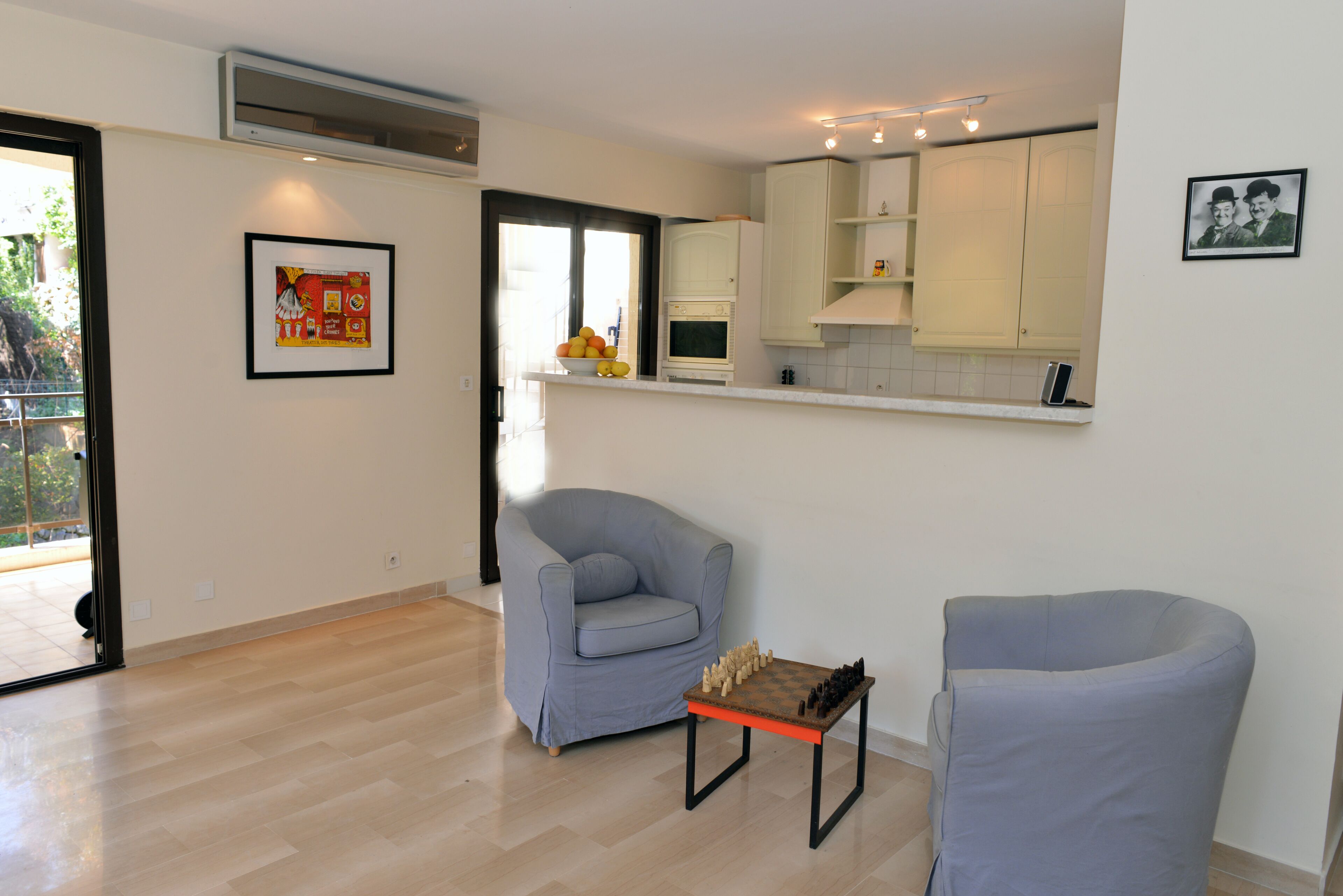 A modern living room with a light blue armchair, a small table with a chessboard, and a kitchen area with white cabinets and a hanging light fixture.