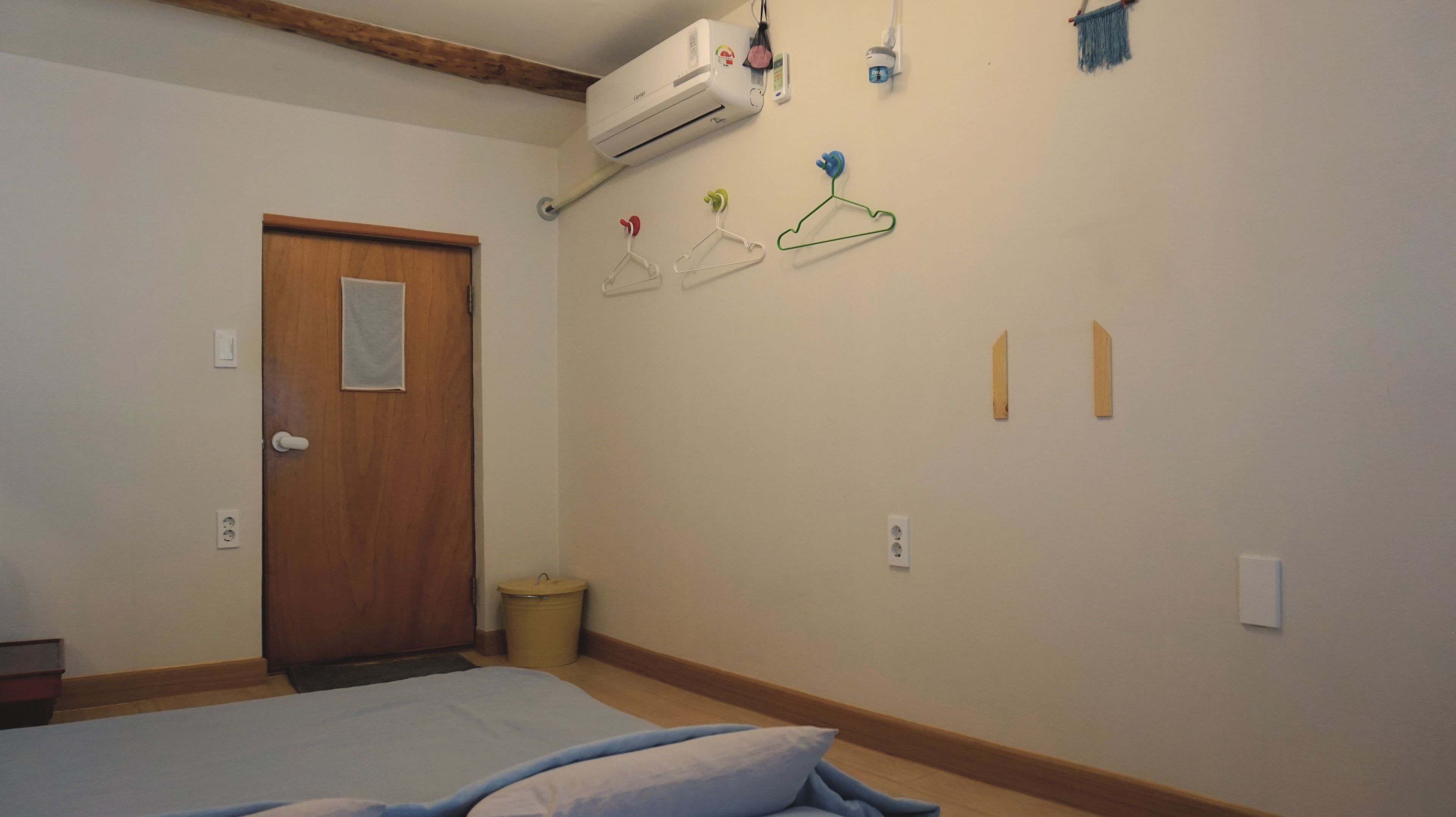 A bedroom with a bed, a wooden door, an air conditioning unit, a small trash bin, and wall-mounted hangers.