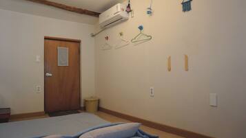 A bedroom with a bed, a wooden door, an air conditioning unit, a small trash bin, and wall-mounted hangers.