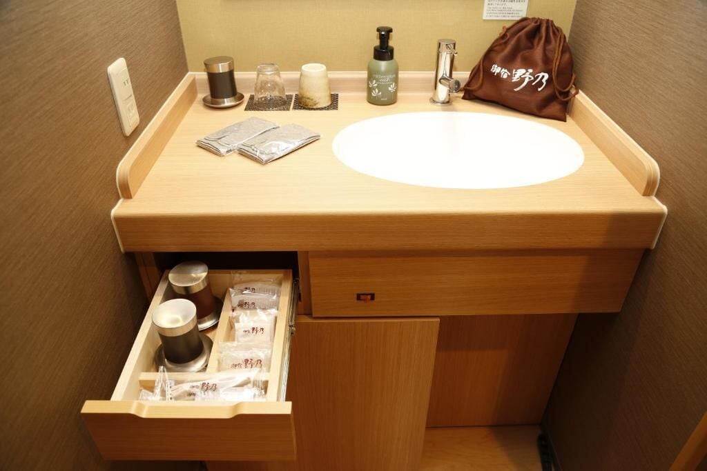 Un baño pequeño con tocador de madera, lavamanos y un cajón con artículos de aseo.
