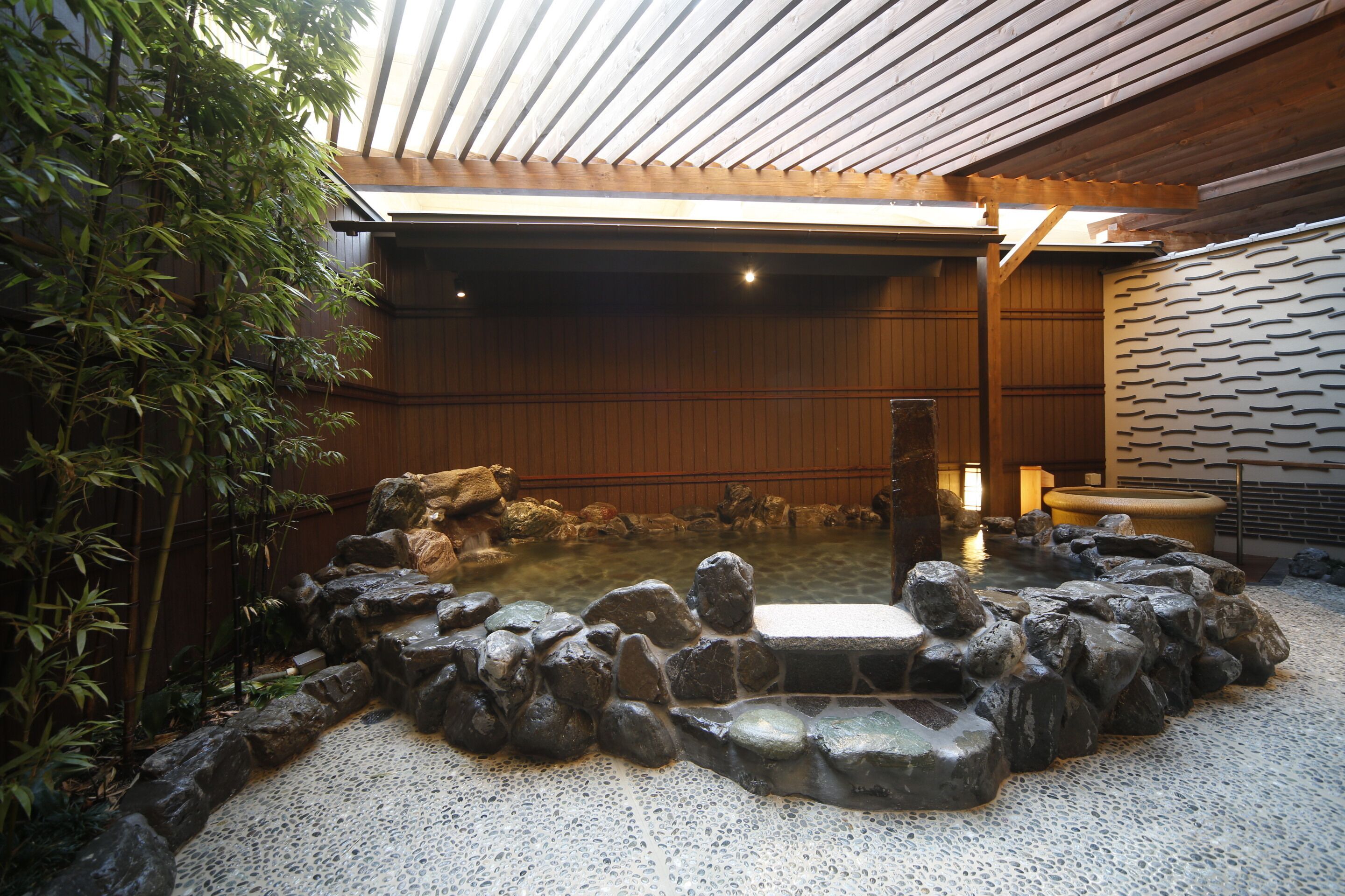Un onsen japonés tradicional con un balneario de piedra rodeado de rocas, paredes de madera y plantas de bambú.