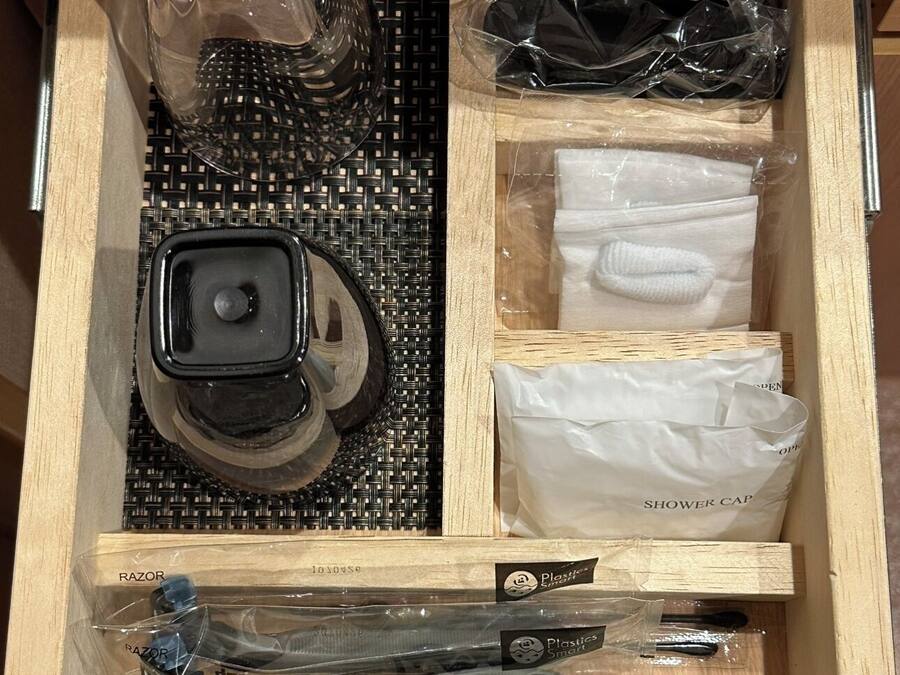 A wooden drawer with compartments containing a razor, a shower cap, a toothbrush, and a plastic bag.