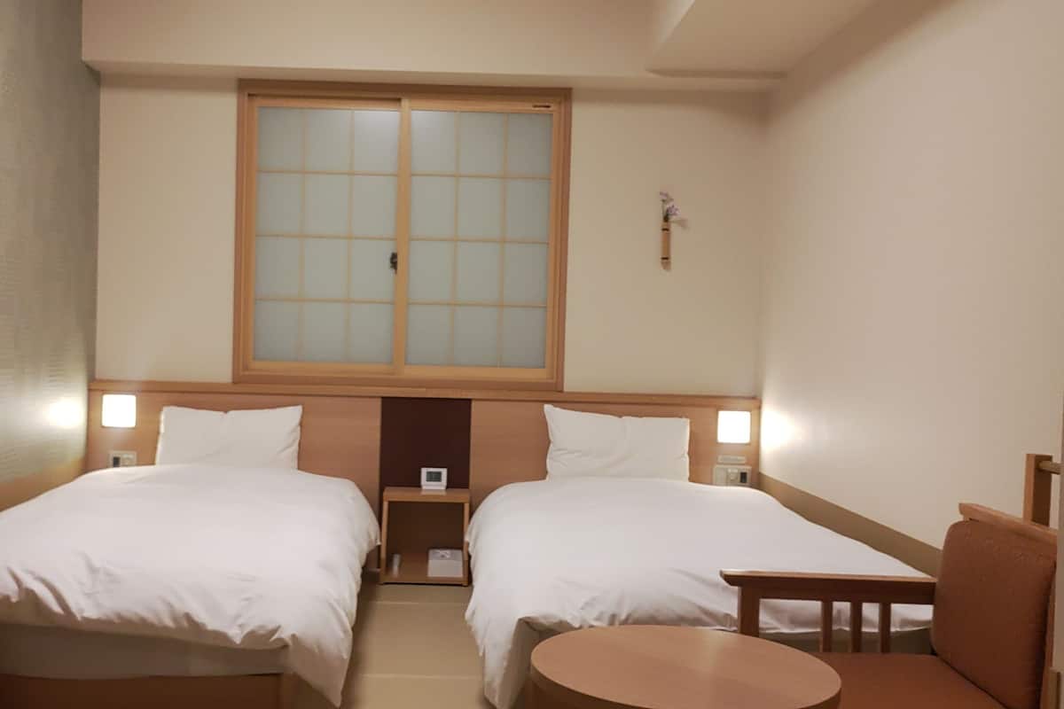 A hotel room with two beds, a small table, and a window with a view.