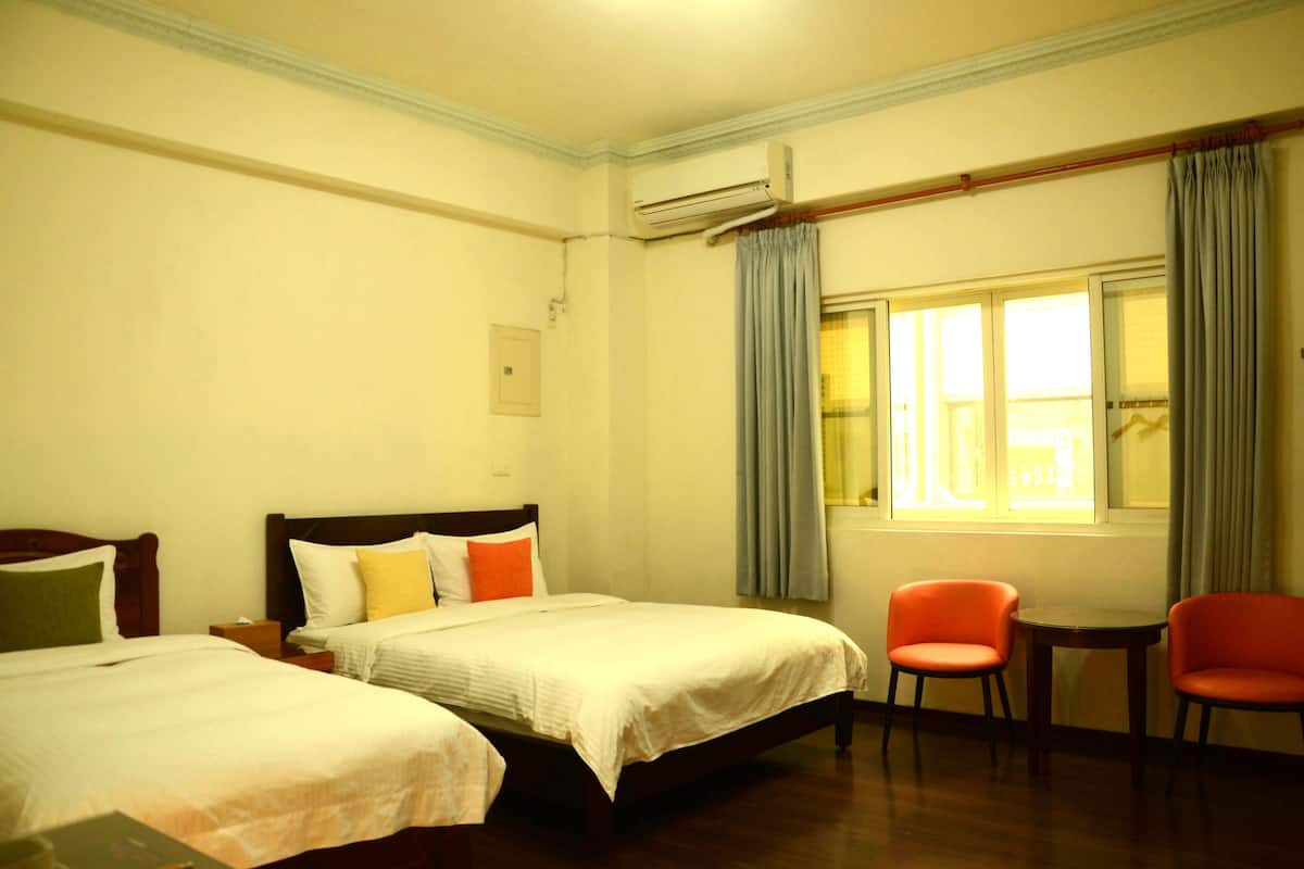Habitación de hotel con dos camas, una mesita pequeña, dos sillas, ventana con cortinas y equipo de aire acondicionado.