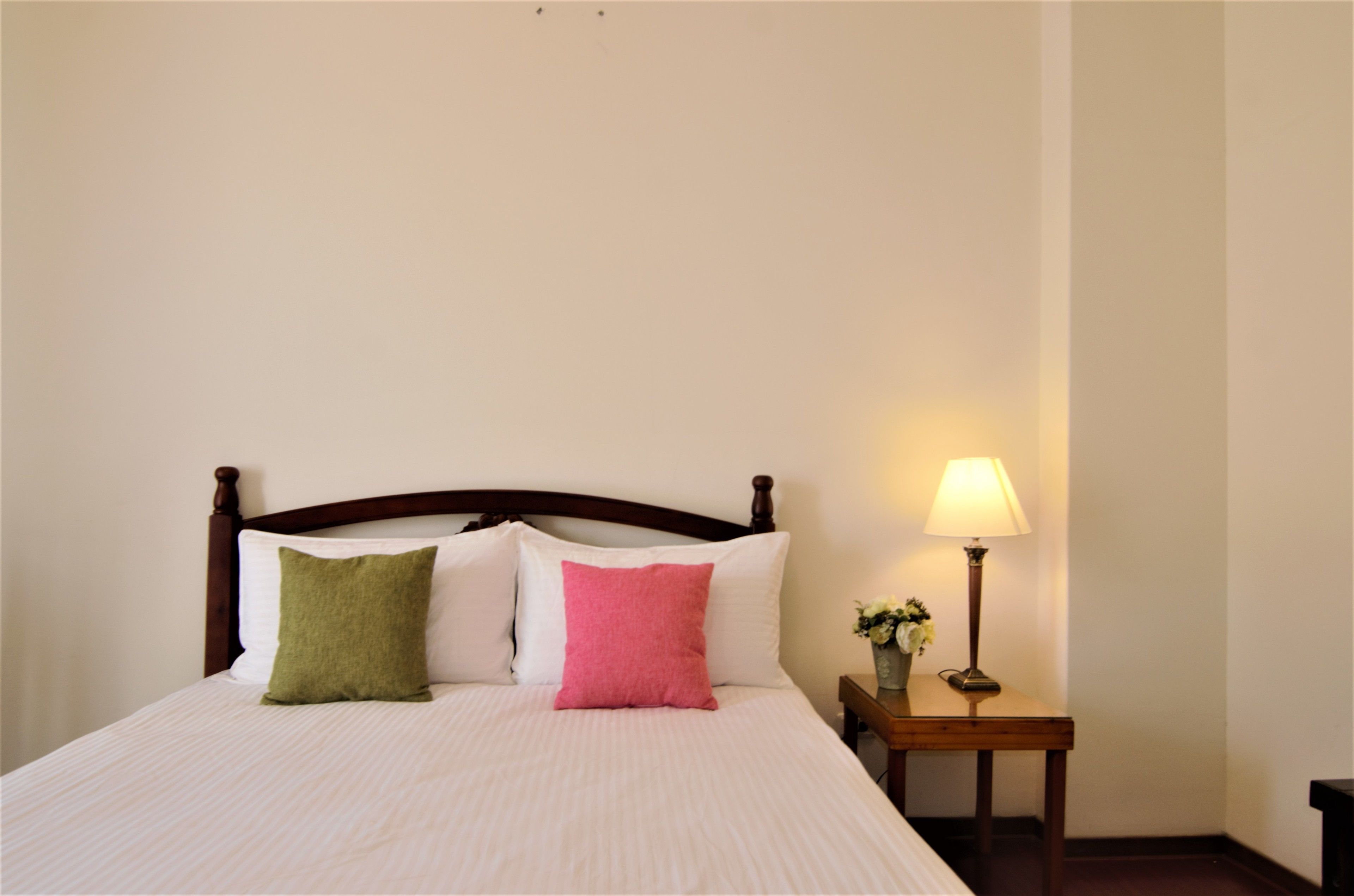 A neatly made bed with a wooden headboard, two pillows, a bedside table with a lamp, and a vase with flowers.