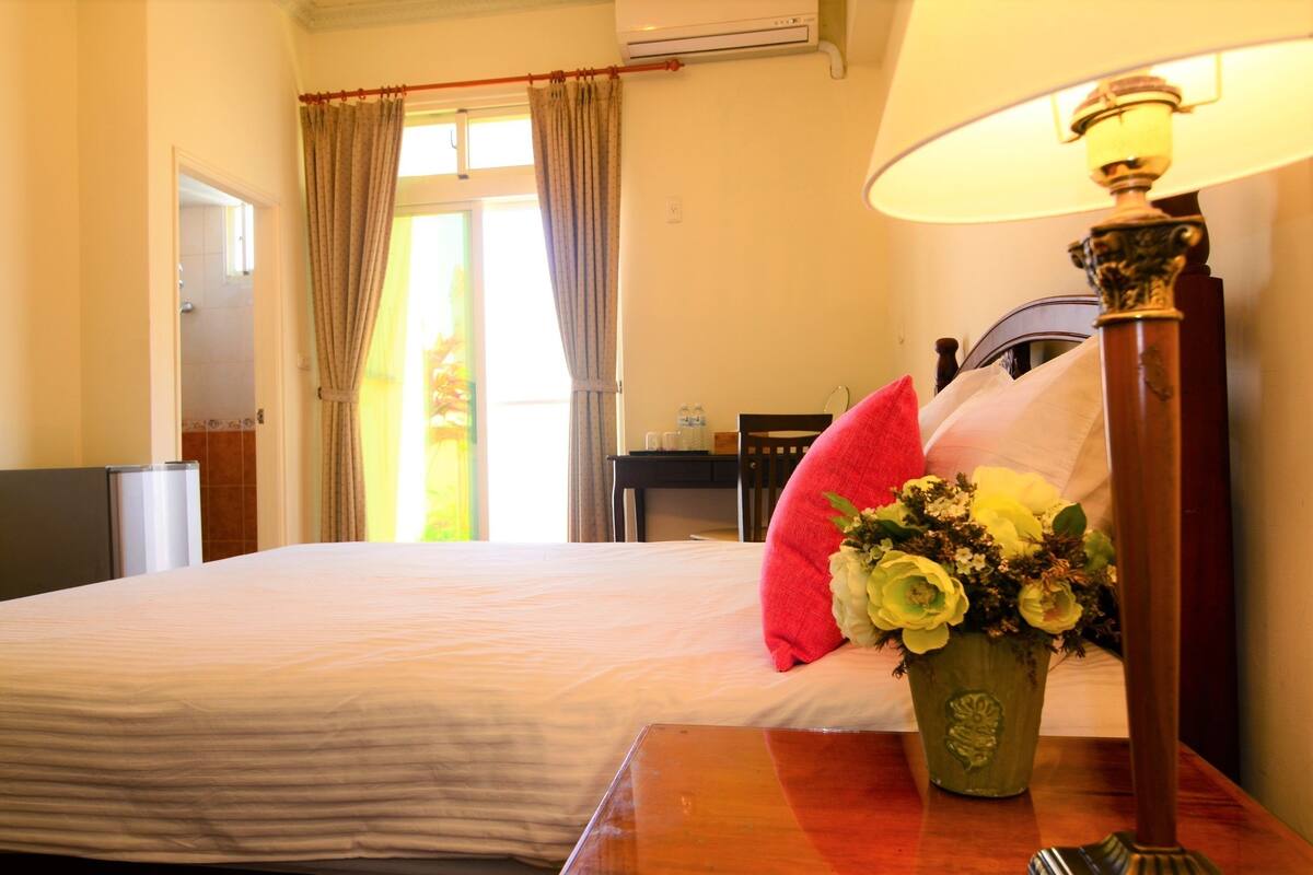 Habitación de hotel con cama, mesita de noche con lámpara, un jarrón de flores y vista al exterior a través de una ventana con cortinas.