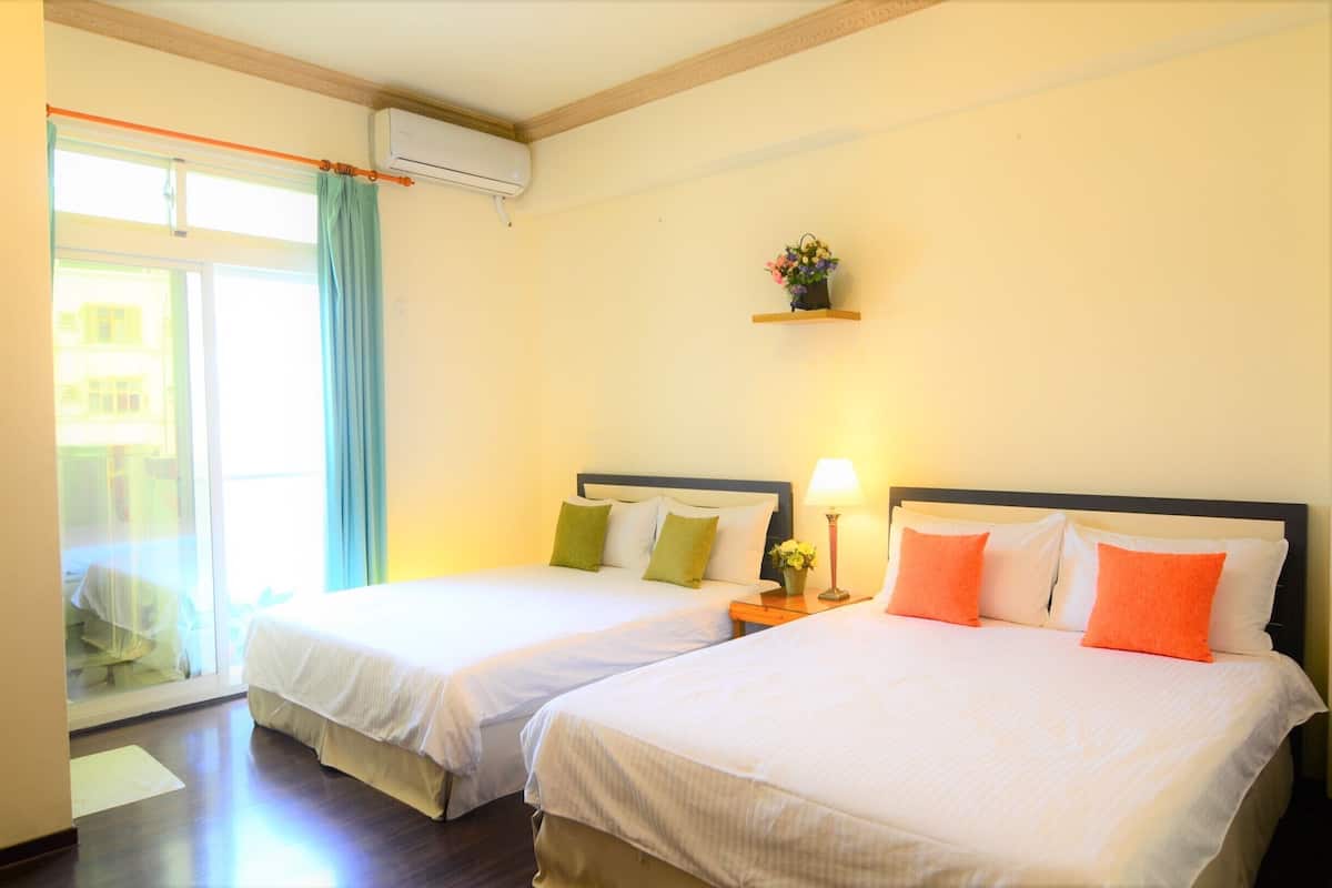 Habitación de hotel con dos camas, una ventana con cortina, unidad de aire acondicionado y un pequeño estante con flores.