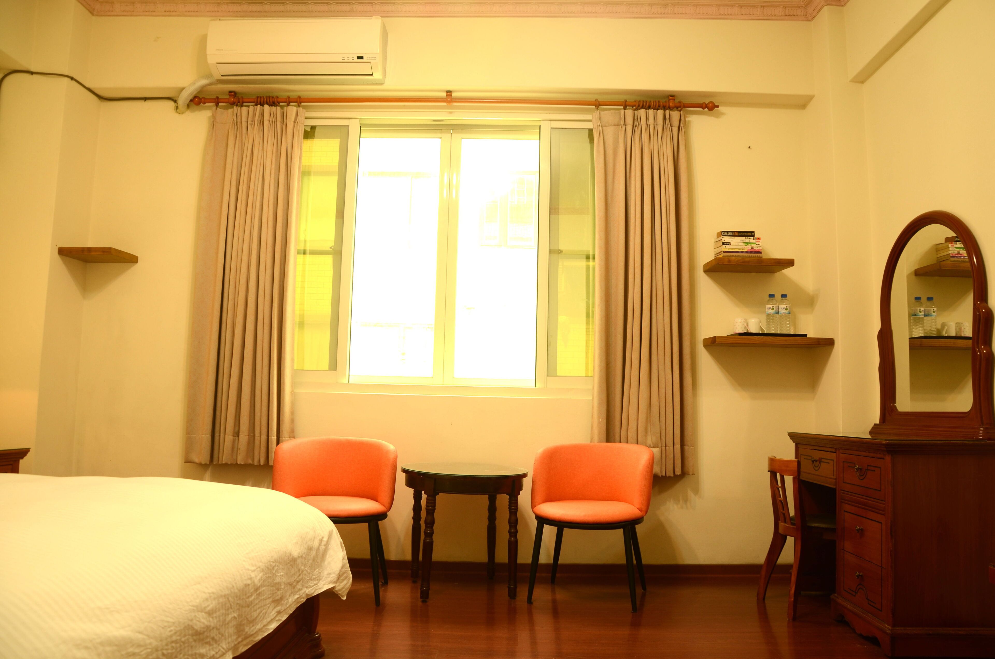 A hotel room with a bed, two orange chairs, a small table, a dresser, and a window with curtains.