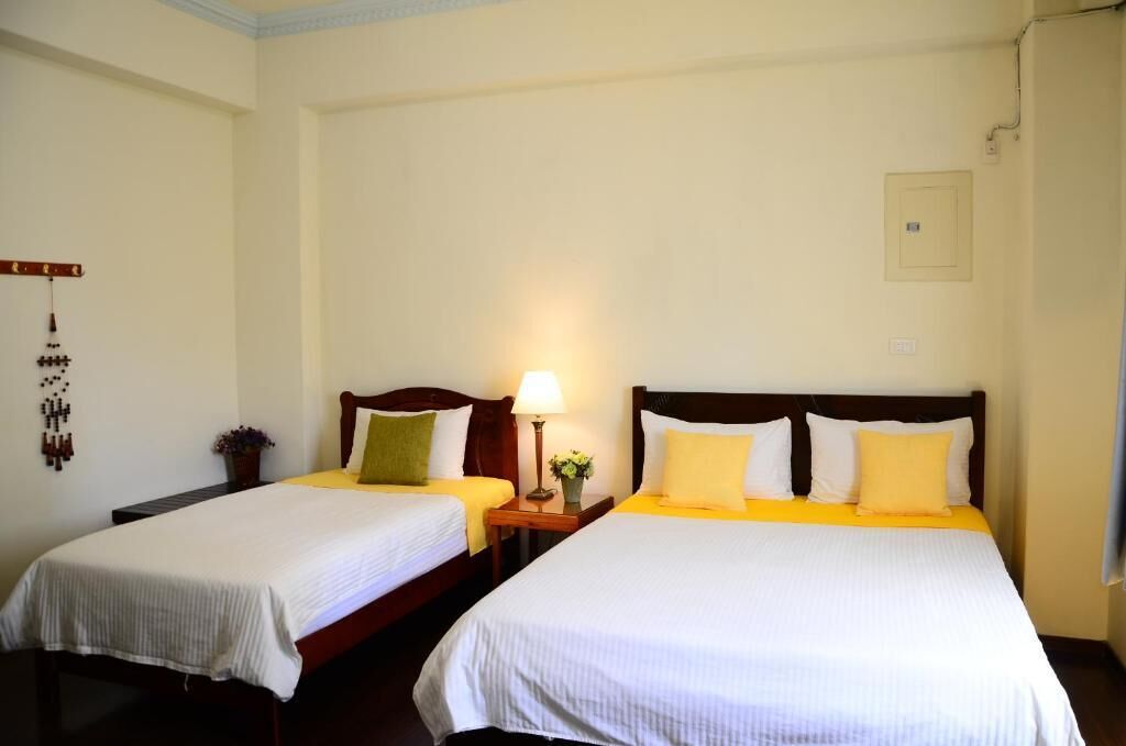 A hotel room with two beds, each with a white bedspread and yellow pillows. There is a nightstand with a lamp and a small plant between the beds.