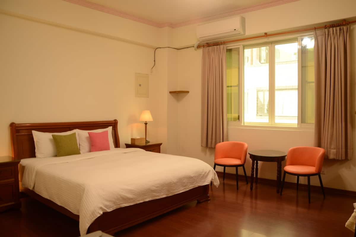 Habitación de hotel con cama, dos sillas, una mesita, equipo de aire acondicionado y ventana con cortinas.